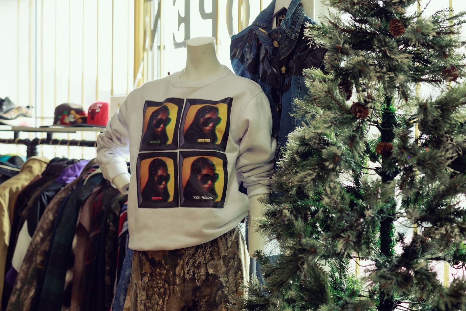 A white crewneck printed with four portraits of Ms. Lauryn Hill on a mannequin beside a Christmas tree